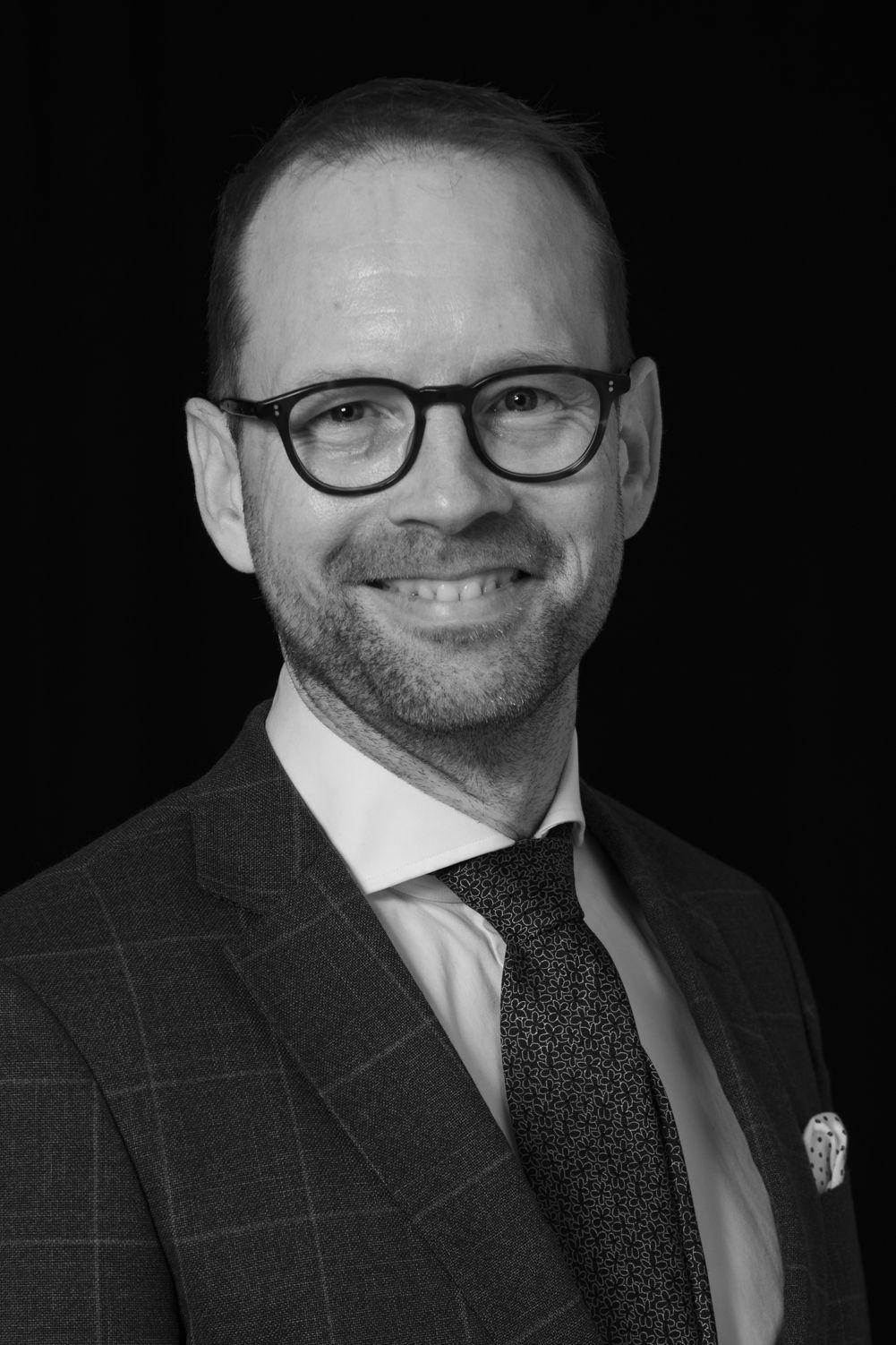 Mustavalkokuva hymyilevästä Harri Leinikasta, silmälaseissa ja tumma puku päällä.