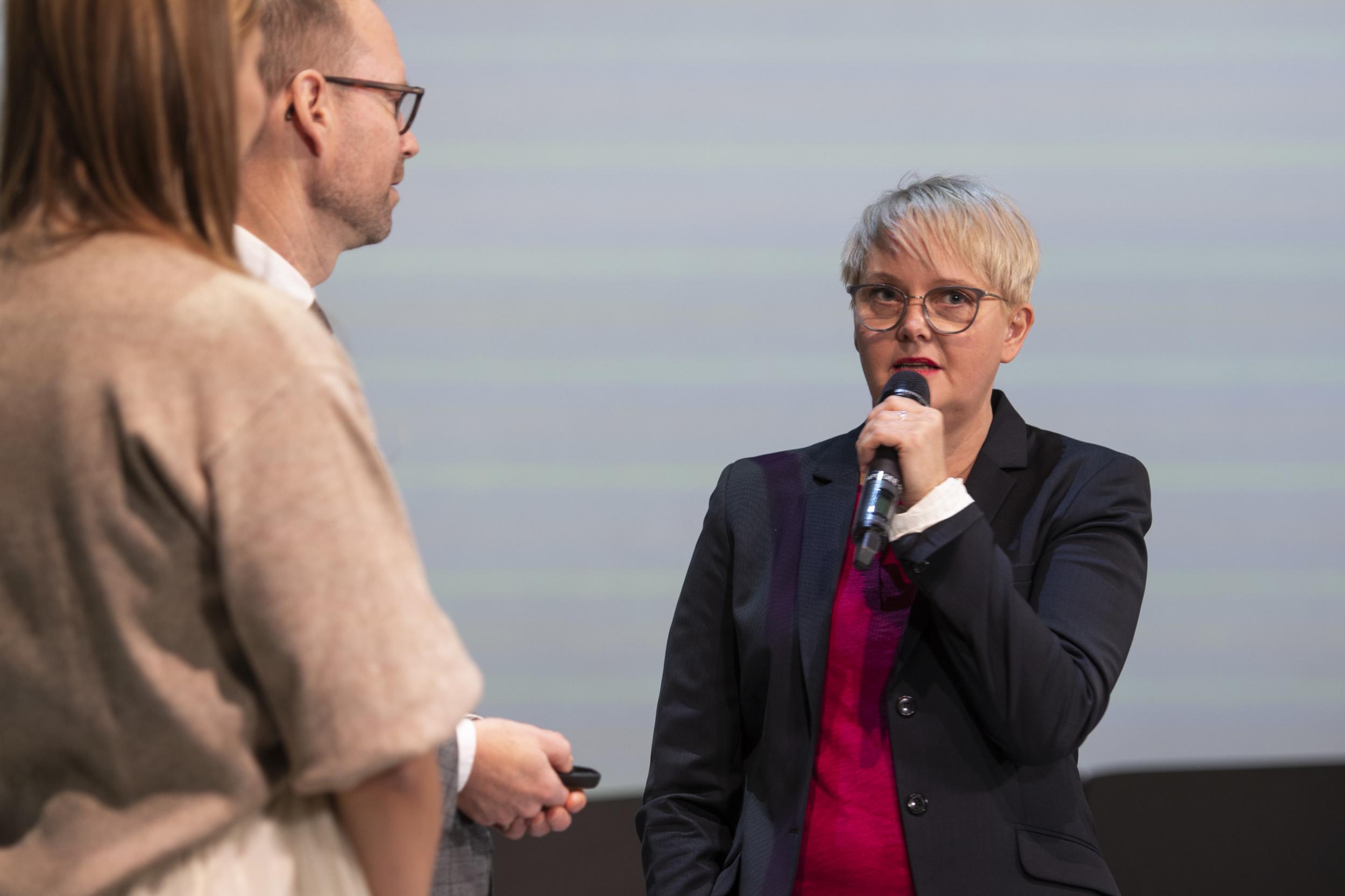 Kaksi haastattelee naista, jolla mikrofoni kädessä.